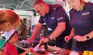 dziecko z opiekunem stojące przy policyjnym stoisku profilaktycznym. Na zdjęciu widać też umundurowanego policjanta i policjantkę