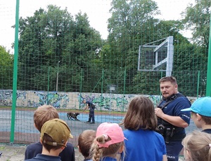 policjant rozmawia z uczniami na boisku szkolnym w tle przewodnik z psem służbowym