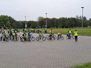 dzieci w kamizelkach odblaskowych i kaskach na rowerach stojące za opiekunem i oczekujące na wyjazd