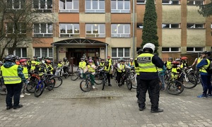 grupa dzieci w kamizelkach i kaskach siedząca na rowerach przed budynkiem szkoły w towarzystwie policjantów