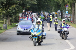 dwoje policjantów jadących na motocyklach za którymi jedzie policyjny radiowóz. W oddali widać maszerujące dzieci oraz policjantów