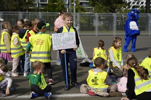 dzieci ubrane w kamizelki odblaskowe na jednej z ulic miasta. Jedno z nich trzyma transparent promujący bezpieczeństwo na drodze. W tle widać policyjną maskotkę