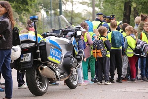 dzieci w kamizelkach odblaskowych ustawione tyłem do zdjęcia stojące obok policyjnego motocykla. W oddali widać funkcjonariuszy straży miejskiej