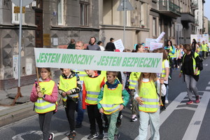 kilkoro dzieci w kamizelach odblaskowych niosące transparent jestem widoczny, jestem bezpieczny. Za dziećmi widać grupę osób biorących udział w marszu