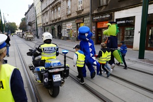 maskotka policji i straży miejskiej prowadzące dzieci w odblaskach przez ulice Krakowa obróceni do zdjęcia tyłem w towarzystwie policjanta na motocyklu