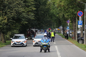 policjant na motocyklu za którym widać pojazdy osobowe mpk, dorożki oraz grupę dzieci oczeukjącą na wymarsz na jednej z krakowskich ulic