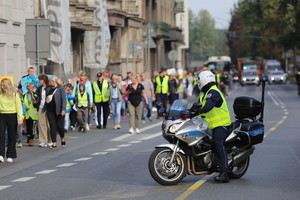 policjant na motocyklu obserwujący grupę dzieci i opiekunów idących w marszu. w oddali widać pojazdy służb