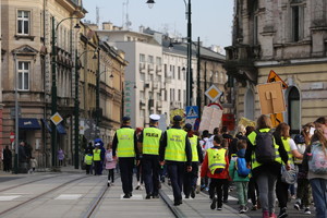 policjant oraz dwóch strażników miejskich maszerują wspólnie z grupą dzieci i opiekunów przez jedna z ulic w centrum miasta. Osoby odwócone do zdjęcia tyłem