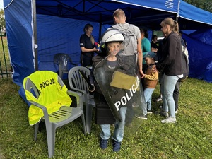 osoby stojące przed stoiskiem profilaktycznym policji. Na pierwszym planie znajduje się chłopiec wyposażony w sprzęt policyjny