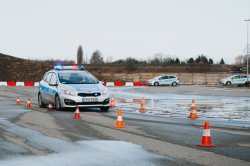 Fotografia przedstawia radiowóz policyjny marki Kia Ceed o numerze rejestracyjnymHPGB-949 podczas wykonywania ćwiczenia „awaryjna zmiana pasa ruchu”.  Widoczny prawy bok i przód samochodu. Radiowóz ma włączone sygnały błyskowe niebieskie i czerwone i porusza się w korytarzu utworzonym z pachołków. Widać przesychające podłoże. W oddali dwa radiowozy czekają na swoją kolej.