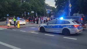 policjant krakowskiej drogówki kieruje ruchem na jednym ze skrzyżowań. Na zdjęciu widać również oznakowany radiwowóz oraz grupę osób oczekujących przed przejściem dla pieszych