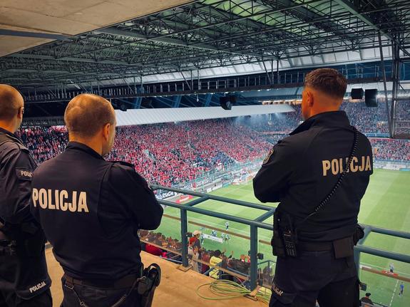 trzech umundurowanych policjantów obserwujących kibiców siedzących na trybunach stadionu