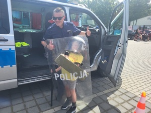 dziecko przymierza policyjny kask i trzyma tarczę obok policjant, w tle radiowóz