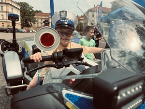 chłopiec trzymając w ręku tarczę do zatrzymywania pojazdów siedzi na policyjnym motocyklu, na głowie ma czapkę policjanta ruchu drogowego