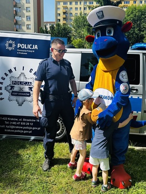 umundurowany policjant w towarzystwie policyjnej maskotki Smoka Bazylego do którego przytula się dwoje małych dzieci