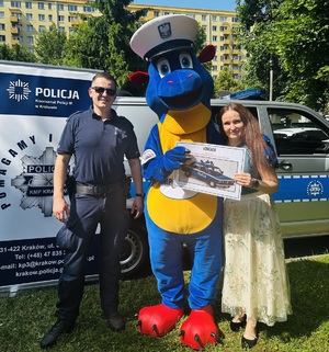 Umundurowany policjant w towarzystwie Smoka Bazylego, który przekazuje voucher przedstawicielce przedszkola