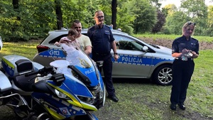 dziecko wraz opiekunem przy policyjnym motocyklu, obok policjant i policjantka, w tle radiowóz