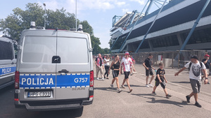 policyjne radiowozy stojące przy stadionie, obok których przechodzą kibice ujdający się na mecz