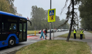 dwie osoby przechodzą przez przejście dla pieszych, przed którym zatrzymał się autobus. Na chodniku stoją dwie policjantki