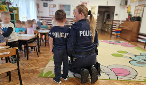chłopiec w koszulce z napisem policja stojący obok umundurowanej policjantki oraz inne dzieci znajdujące się w klasie