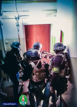 Policjanci wyprowadzają mężczyznę z założonymi kajdankami na ręce i nogi z budynku lotniska.