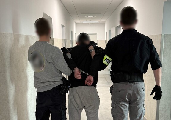 dwóch nieumundurowanych policjantów prowadzi zatrzymanego z kajdankami założonymi na ręce trzymane z tyłu