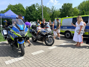 dzieci siedzące na policyjnych motocyklach. Obok widać umundurowanych policjantów oraz osoby dorosłe. Na zdjęciu widać również oznakowany radiowóz oraz stoisko profilaktyczne