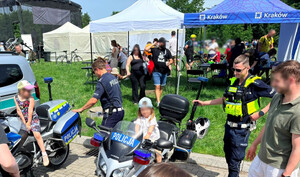 małe dzieci siedzące na policyjnych motocyklach przy których znajdują się umundurowani policjanci. Na zdjęciu widać również stosika oraz osoby zainteresowane atrakcjami
