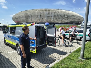 umundurowany policjant stojący przy oznakowanym radiowozie . Obok widać dwóch rowerzystów. W tle widać krakowską Tauron Arene