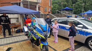 dziecko siedzące na policyjnym motocyklu obok przy radiowozie policjantka i dwoje dzieci, w tle policjant i ratowniczka medyczna