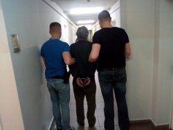 dwóch policjantów kryminalnych prowadzi zatrzymanego mężczyznę, który ma założone kajdanki na ręce trzymane z tyłu