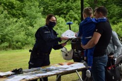 przy policyjnym stoisku profilaktycznym stoi mężczyzna z dzieckiem na ręku, policjantka wręcza dziecku kolorowanki