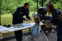 przy policyjnym stoisku profilaktycznym policjantka wręcza dziewczynce opaskę odblaskową obok stoi kobieta, w tle policyjny motocykl