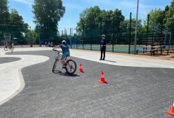 Dziecko w trakcie egzaminu praktycznego na kartę rowerową poruszające się na rowerze po torze przeszkód między pachołkami, w tle policjantka Wydziału Ruchu Drogowego KMP Kraków.