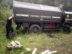 Policyjny przewodnik psa służbowego wraz z psem w trakcie działań na poligonie, obok duży pojazd patrolu saperskiego