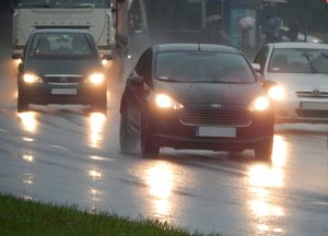 Perspektywa jezdni o trzech pasach ruchu w jednym kierunku, po której jadą (na wszystkich tych pasach) samochody z włączonymi światłami mijania. Przejrzystość powietrza jest obniżona – pada deszcz. Jezdnia jest mokra i śliska – odbija światła reflektorów.