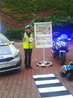 policjantka na dziedzińcu przedszkola w otoczeniu radiowozu policyjnego motocykla oraz tablicy edukacyjnej trzymając znak drogowy wyjaśnia jego znaczenie