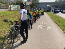 policjant na ścieżce pieszo rowerowej stoi obok służbowego roweru i obserwuje będąca na rowerach grupę dzieci