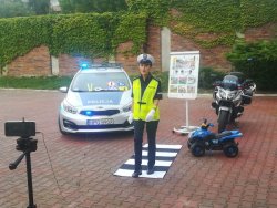 policjantka na_dziedzińcu przedszkola na tle radiowozu i policyjnego motocykla prezentuje jak właściwie  przechodzić przez przejście dla pieszych wykorzystuje do tego matę w formie pasów_0