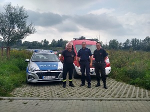 wspólne zdjęcie strażaków i policjanta na tle zalewu i samochodów służbowych