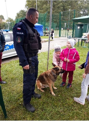 przewodnik psa służbowego rozmawia z dziećmi o swojej pracy. obok policjanta i dzieci siedzi pies