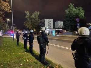 Kilku policjantów stoi w umundurowaniu służbowym przy jedenej z ulic, w tle widać policyjne radiowozy na sygnałach