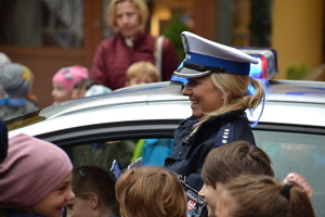 Policjantka ruchu drogowego prowadzi prelekcję stojąc przy radiowozie. W tle otaczające ją dzieci