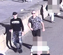 dwóch mężczyzn idących wzdłuż muru na bulwarach wiślanych. Jeden ubrany w czarną bluzę z białym napisem na przodzie oraz szare spodnie, na głowie czarna czapka. Drugi z mężczyzn ubrany w koszulkę w biało szary wzór i czarne spodnie nieokreślonej długości
