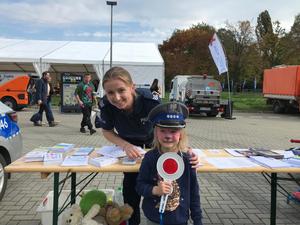 dziewczynka w policyjnej czapce z tarczą do zatrzymywania pojazdów, za nią policjantka w tle ludzie i samochód cysterna