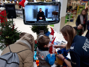 policjantka dziecko i kobieta stoją tyłem. Na wprost monitor wyświetlający przemawiającego policjanta. w tle wnętrze marketu i ludzie