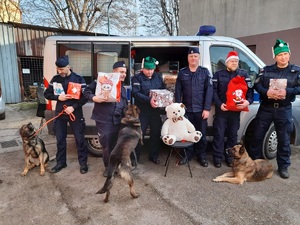 policjanci oraz psy służbowe przy radiowozie. policjanci trzymają precenty dla osoby, którą mają obdarować