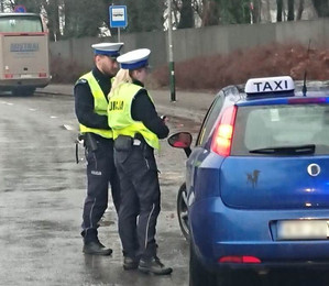 policjant i policjantka stojący obok taksówki, prowadzą rozmowę z kierowcą znajdującym się w pojeździe