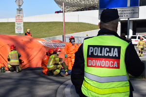 dowodzący działaniami policjant stojący tyłem do zdjęcia. Na dalszym planie widać inne słyzby udzialające pomocy osobie poszkodowanej