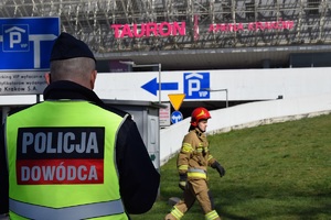 dowodzący działaniami policjant stojący tyłem do zdjęcia. Na dalszym planie widać idącego strażaka